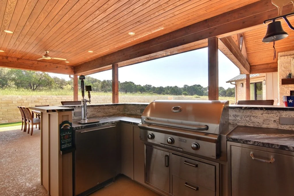 Outdoor kitchen with stainless steel grill, granite counters, and a view of a backyard with trees and a clear sky.