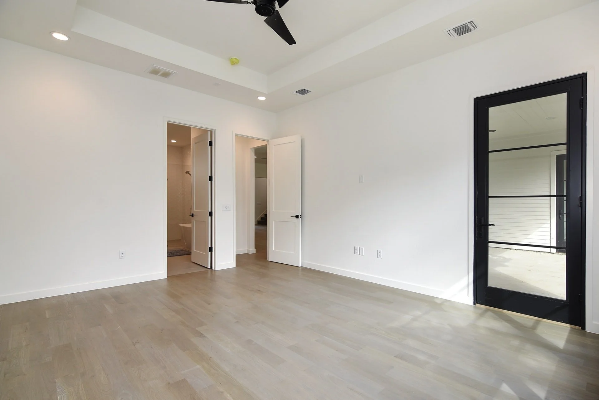 Empty room with light wood flooring, white walls, and a black-framed glass door leading outside. There are two open white doors leading to other rooms and a ceiling fan with black blades.