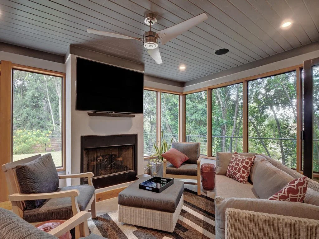 Living room with large windows showing trees, fireplace, wall-mounted TV, ceiling fan, sofa with throw pillows, armchairs, and ottoman.