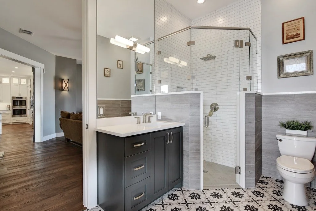 Modern bathroom with a walk-in shower with glass doors, a black vanity with a white countertop, and a toilet topped with a plant. The floor has black and white patterned tiles, and the walls are decorated with framed pictures.