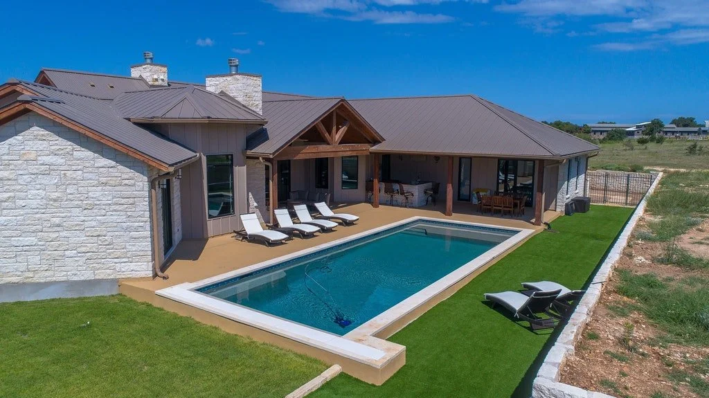 A backyard with a rectangular swimming pool, surrounded by a green lawn and a concrete patio, with lounge chairs and a house with a brown roof and stone exterior.