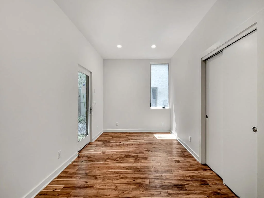 Empty room with white walls, hardwood floors, a glass door leading outside, a window facing a neighboring building, and a sliding closet door.