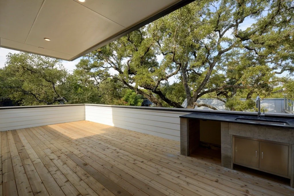 Empty outdoor balcony with wooden decking, white railing, and small outdoor sink with cabinet, overlooking trees.