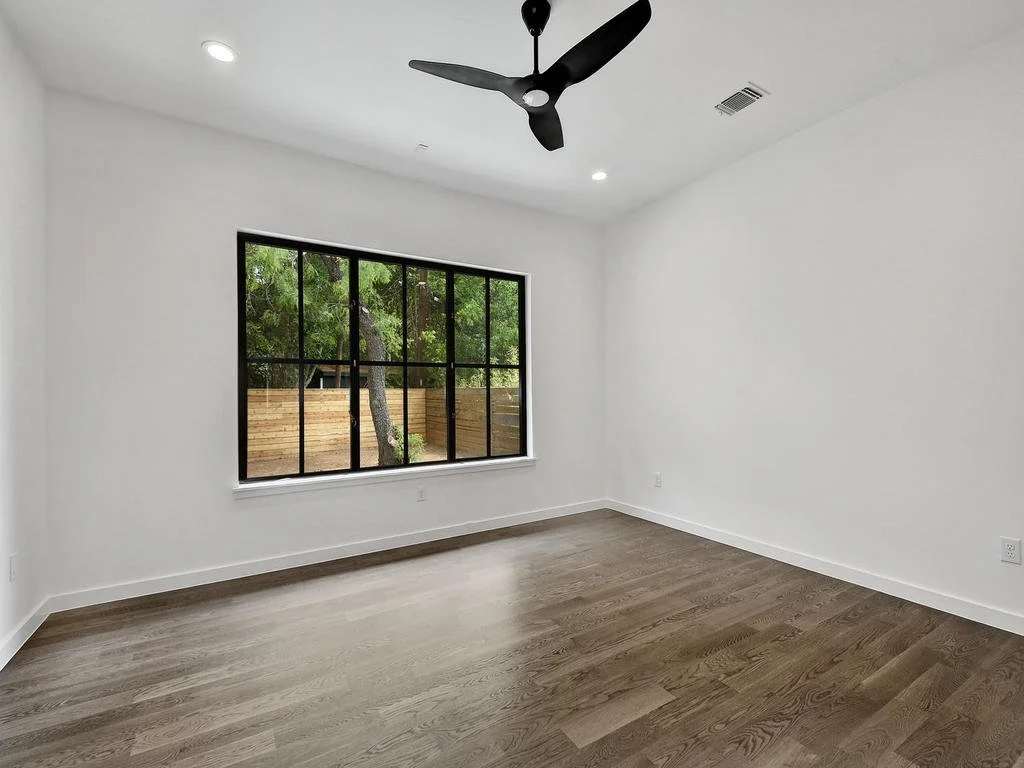 Empty room with white walls, wooden floor, a large window showing trees and a wooden fence outside, a black ceiling fan, and recessed lighting.