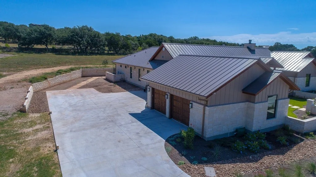 Newly built house with metal roof, attached garage, concrete driveway, and landscaped yard, situated in a rural area with trees and open fields.