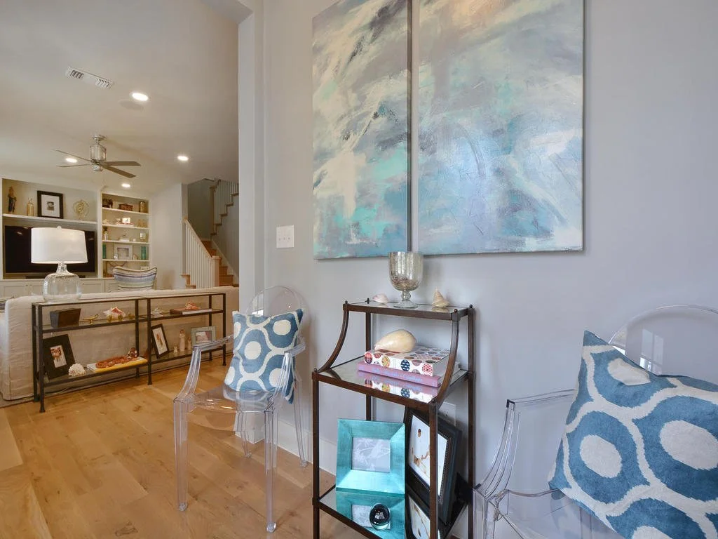 Living room with light wood flooring, a beige couch, a glass and metal side table with decorative items, a large abstract painting on the wall, and transparent chairs with blue patterned cushions. There is a ceiling fan and recessed lighting.