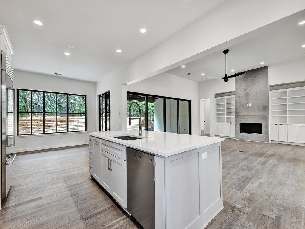 Modern open-concept kitchen and living room with large windows, a fireplace, built-in white shelves, and a ceiling fan.