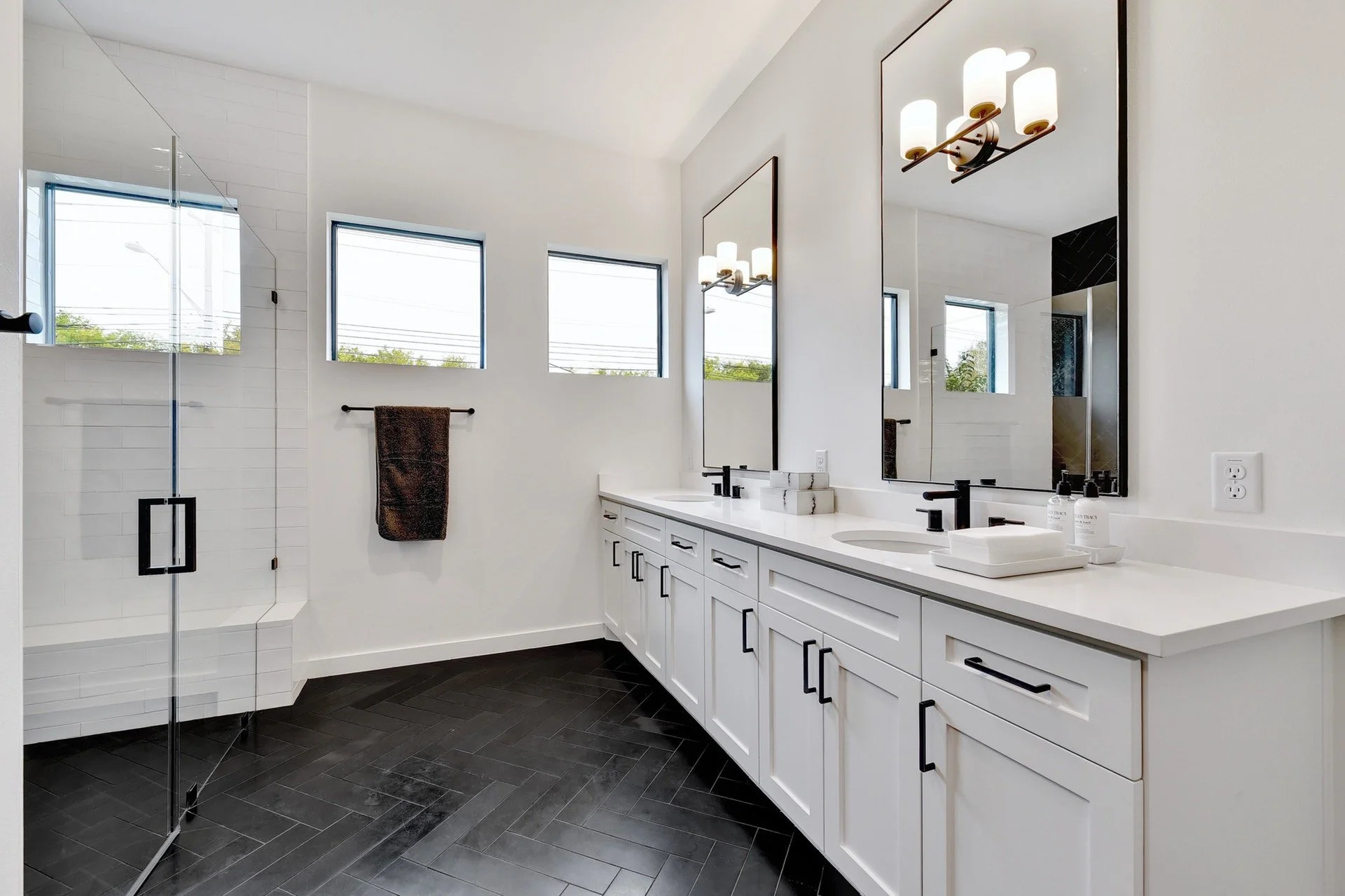 Modern bathroom with a double vanity, large mirrors, black fixtures, and a walk-in shower with glass door, white walls, and three windows.