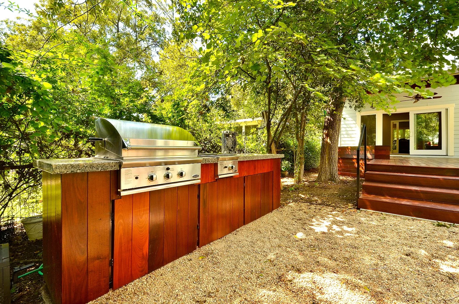 An outdoor grilling station with a stainless steel gas grill on a wooden counter, surrounded by trees and bushes, next to a house with a wooden deck.