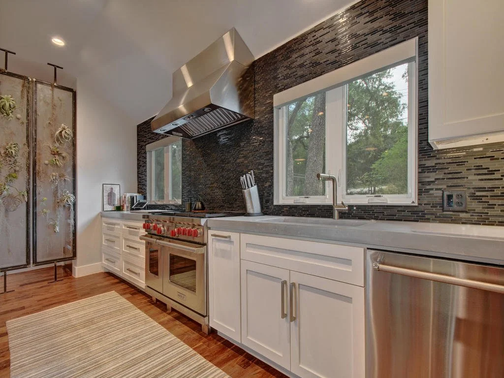 Modern kitchen with white cabinets, a black tiled backsplash, stainless steel appliances, a large window, and hardwood floors.