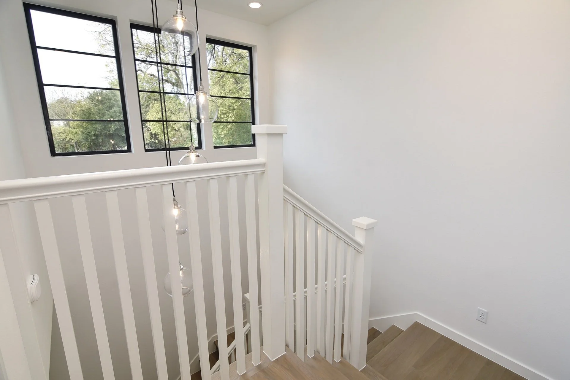 Interior view of a staircase landing with white wall, black-framed windows showing trees outside, and hanging glass pendant lights.