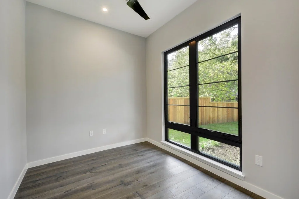 Empty room with gray walls, hardwood flooring, large window showing a wooden fence and greenery outside, ceiling fan, and recessed lighting.