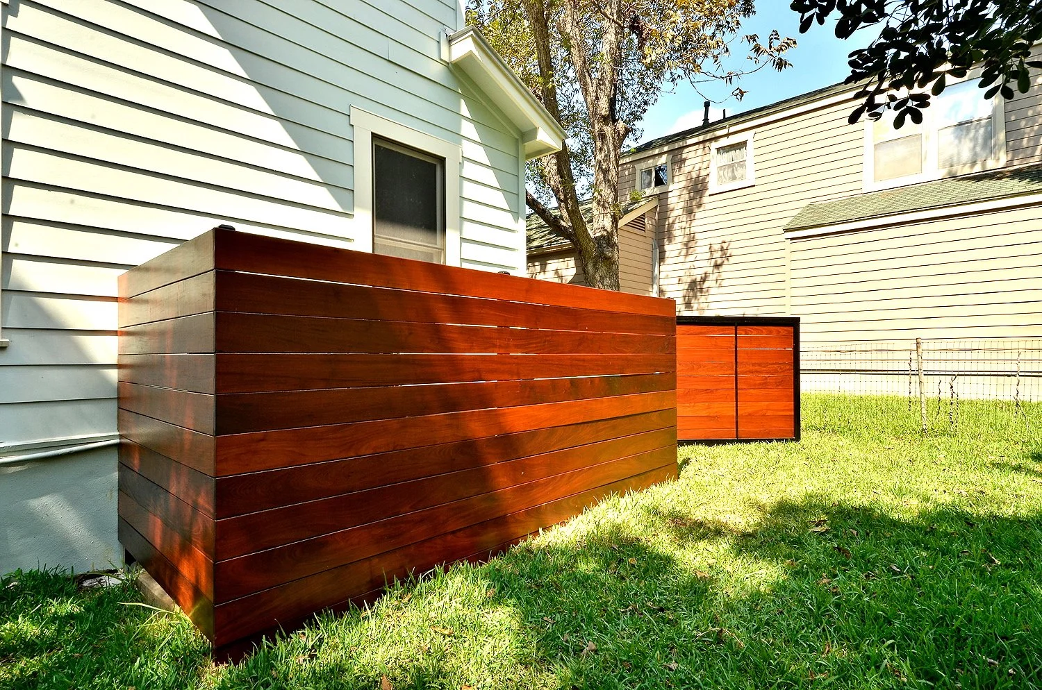 Close-up of modern wooden fence panels with horizontal planks, installed along the side of a white house with vinyl siding, in a backyard with green grass and trees.