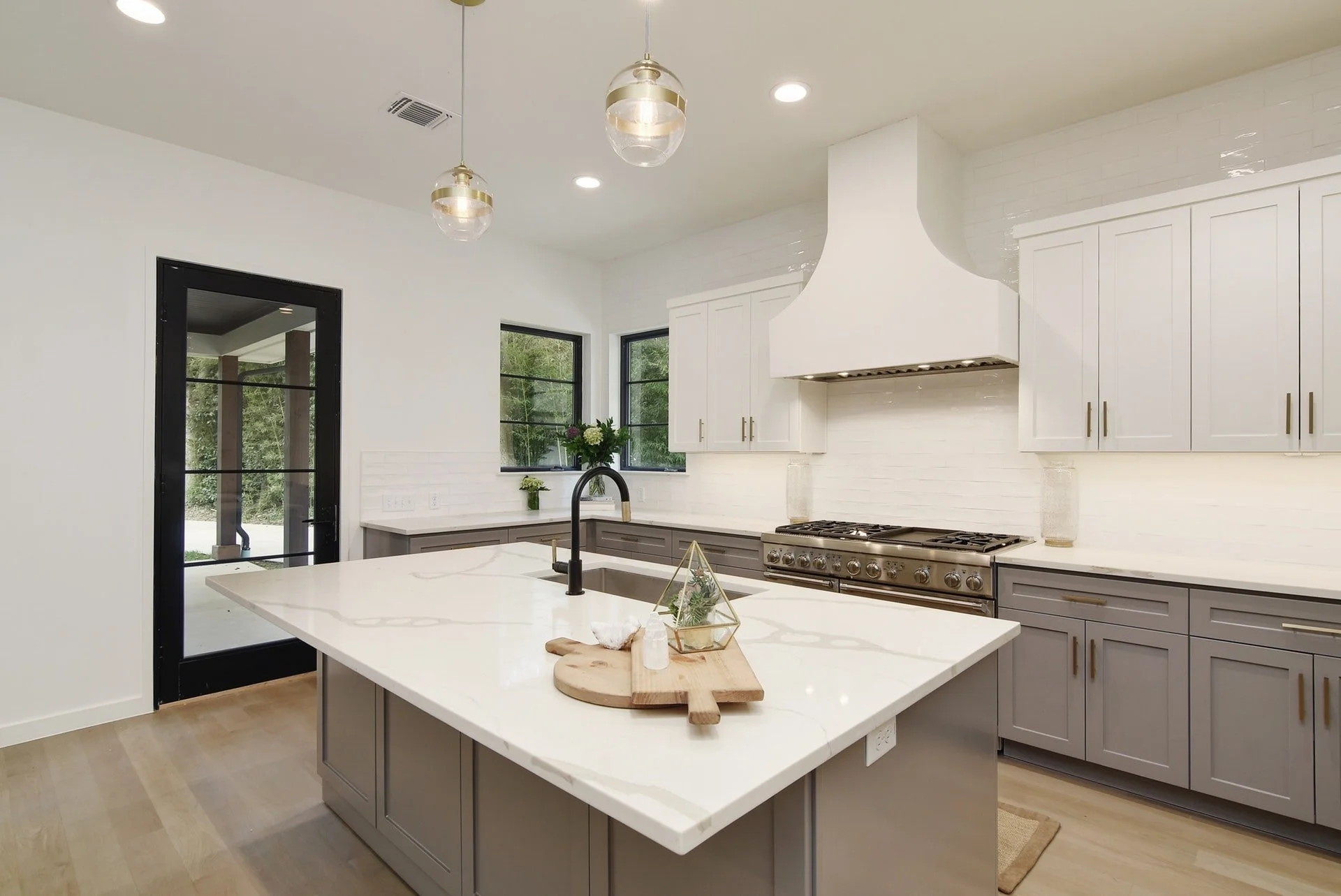 Modern kitchen with white walls, white upper cabinets, gray lower cabinets, and a large white island with a built-in sink and black faucet. Stainless steel stove and range hood, two pendant lights, two windows, and a glass door leading outside. Decor