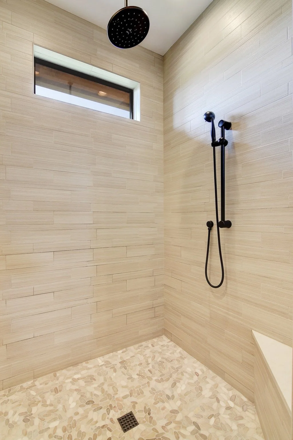 A modern shower with beige tiled walls, pebble stone floor, a black rainfall showerhead from the ceiling, a black handheld shower, a small window high on the wall, and a corner seat.
