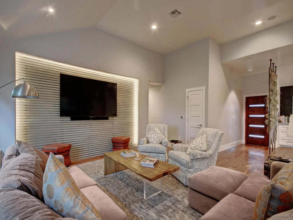 A cozy living room with a large TV mounted on a wall with horizontal slats. The room has a beige sofa, patterned armchairs, and small red stools. A wooden coffee table with magazines is centered, and there's a floor lamp to the left.