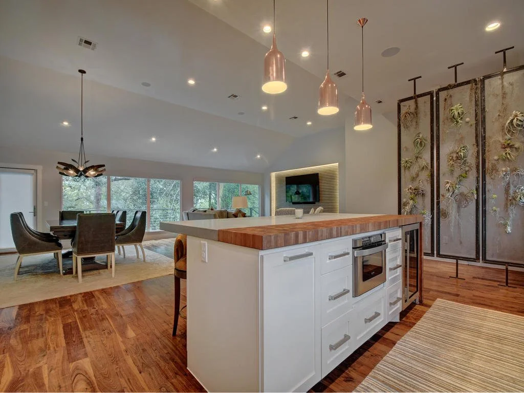 Open-concept living and dining space with hardwood floors, a kitchen island with white cabinets and a wooden countertop, modern pendant lights, a dining table with six chairs, a wall-mounted TV, and a decorative vertical garden wall.