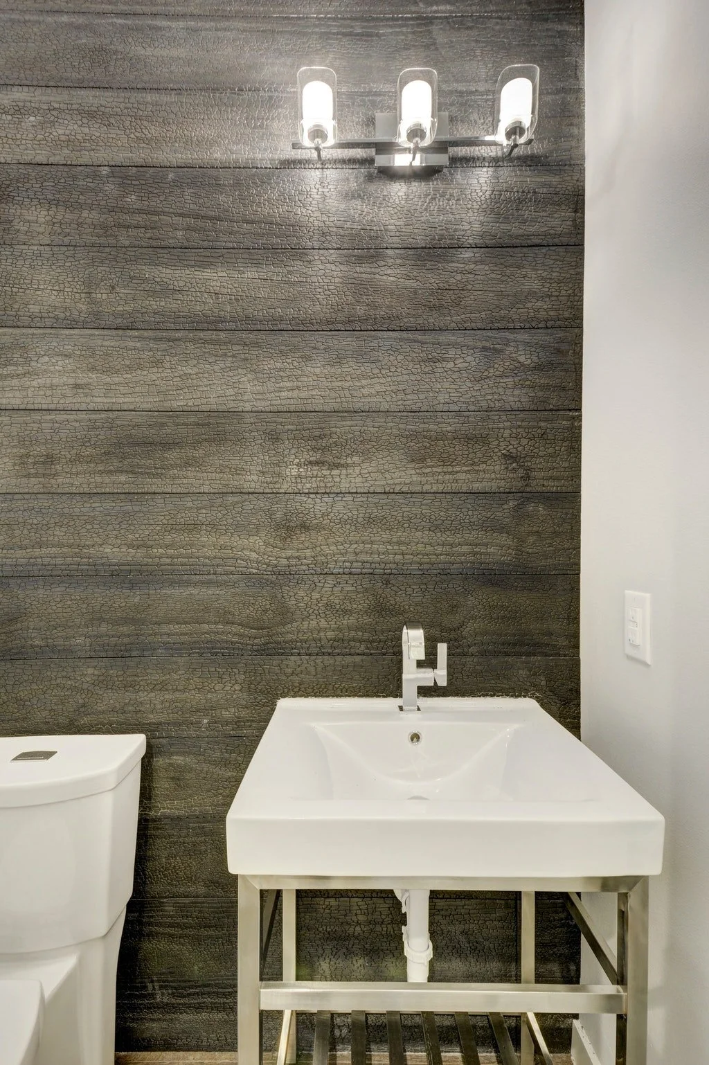 A modern bathroom with a white toilet, a white sink on a metal frame, a dark textured wood wall, and a three-light fixture above.