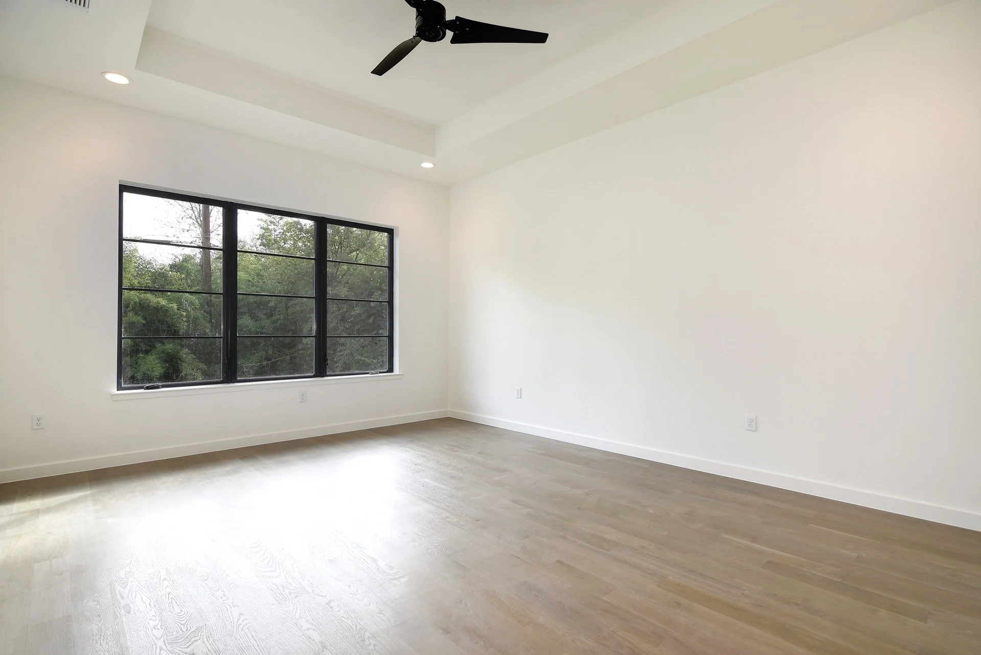 Empty room with large window, white walls, light wooden floor, ceiling fan, and recessed lighting.