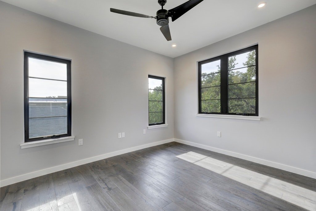 An empty room with three black-framed windows, gray walls, hardwood flooring, a black ceiling fan, and recessed lighting.