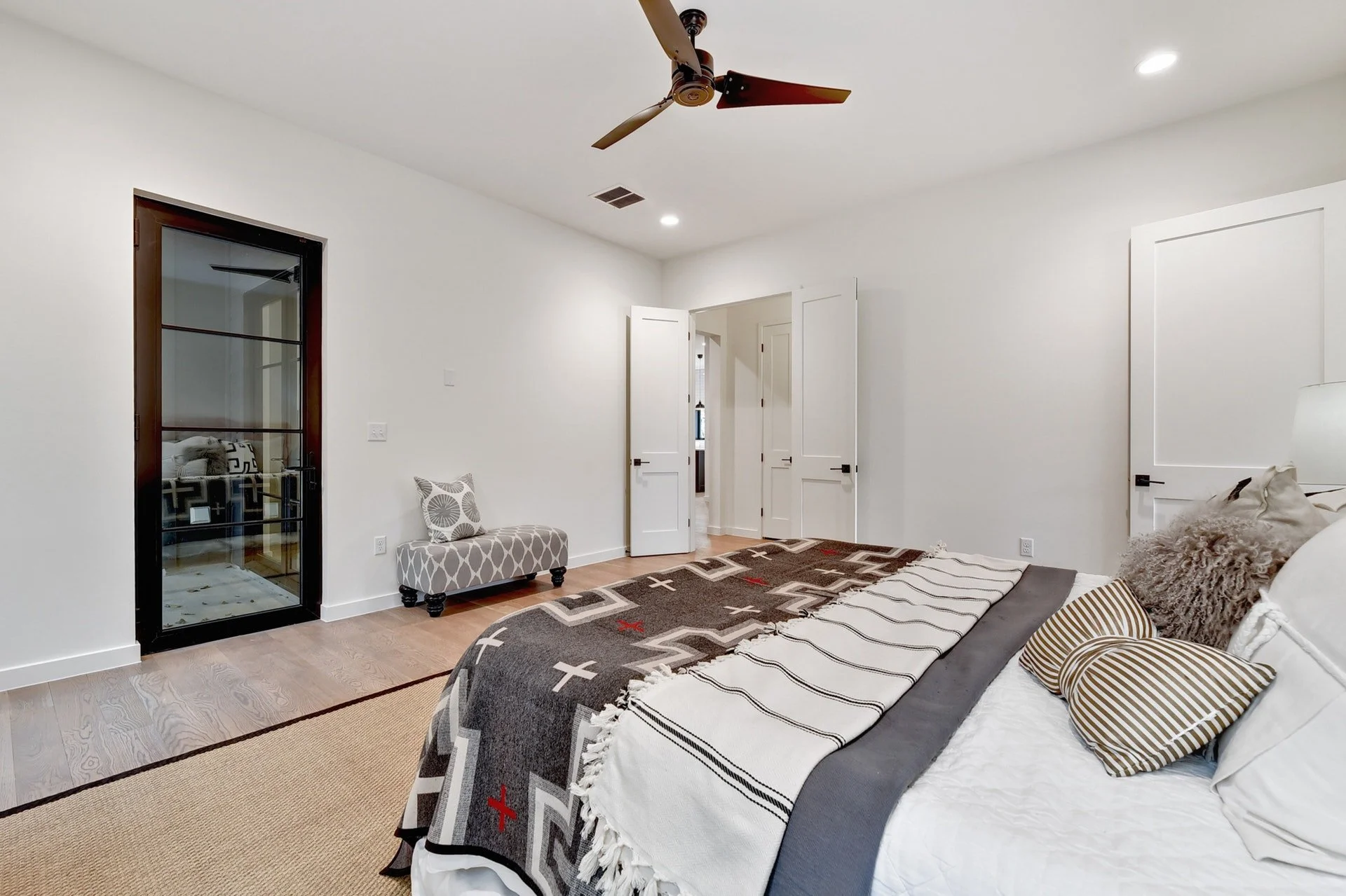 A bedroom with a bed covered in a black, white, and gray geometric patterned quilt and decorative pillows, including a fluffy gray pillow and a striped pillow. There is a small upholstered bench with a gray and white pattern near a white wall with a 