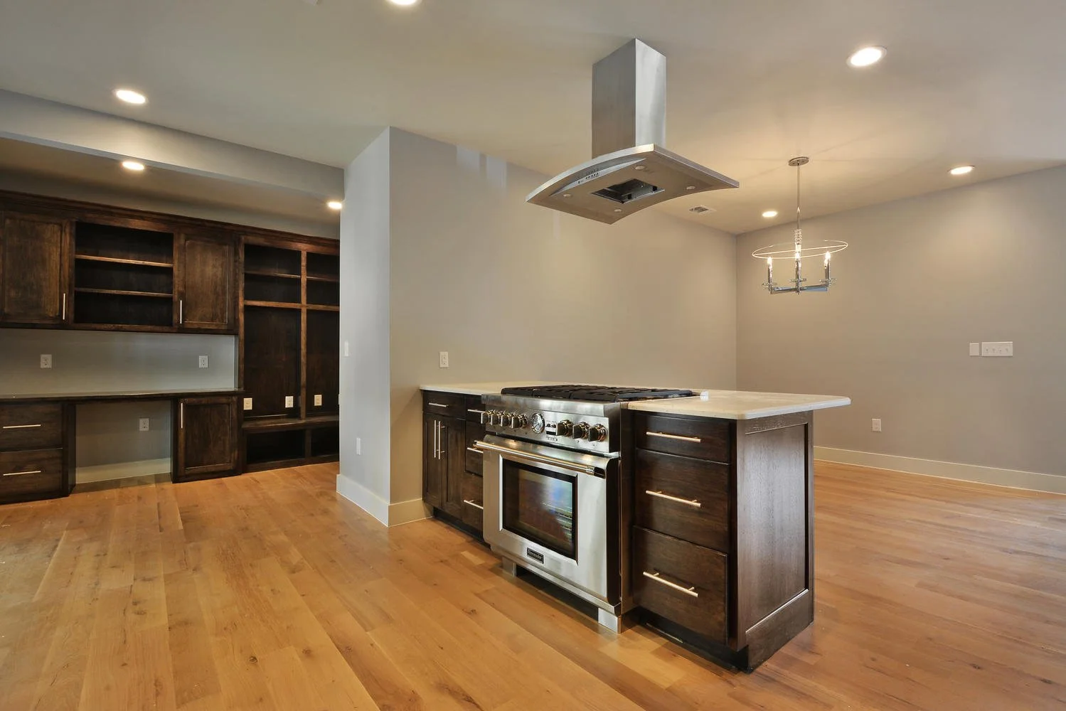 Empty kitchen with dark cabinets, an island with a stove, a stainless steel range hood, and hardwood floors.