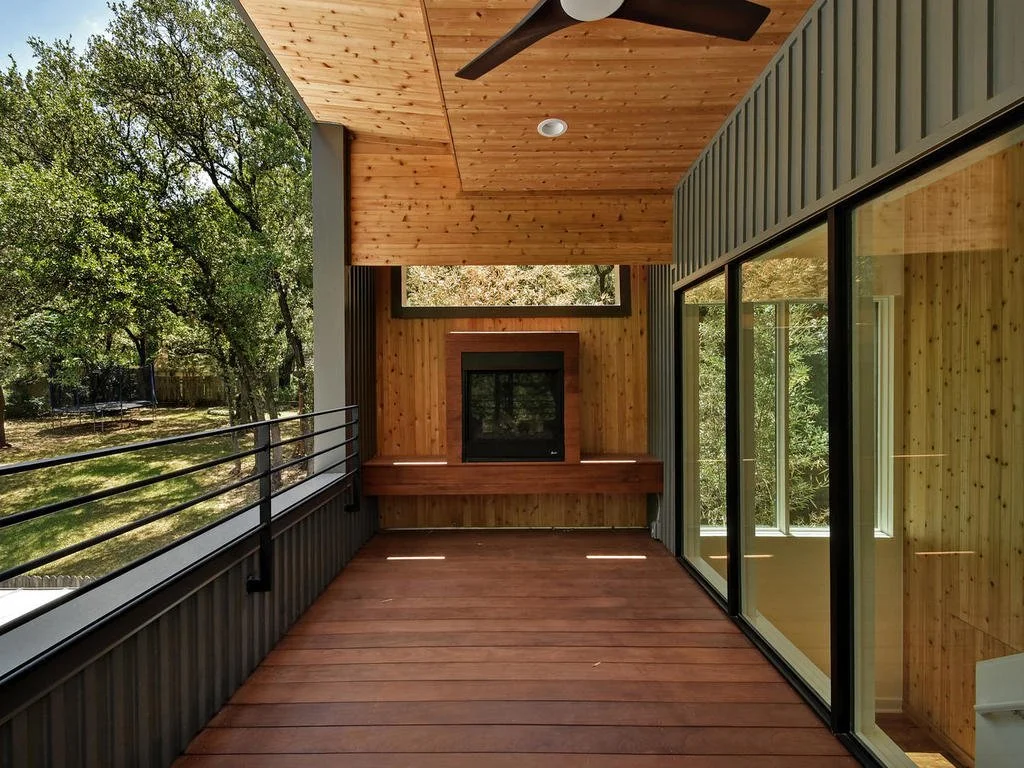 A covered balcony with wooden flooring, wood-paneled ceiling, large sliding glass doors, and a fireplace with a wooden mantel, overlooking trees and greenery.