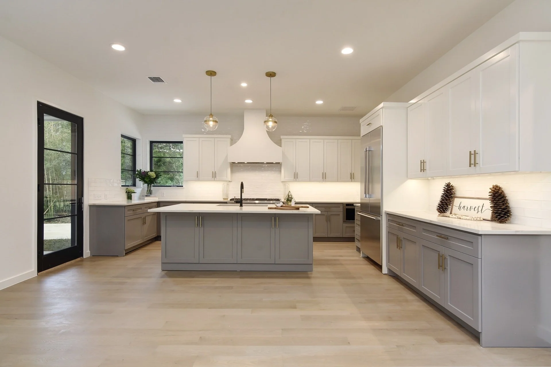 Modern kitchen with white walls and ceiling, gray cabinets, white countertops, and hardwood flooring. Features include kitchen island, pendant lighting, stainless steel appliances, and decorative items like pinecones and a 'harvest' sign.