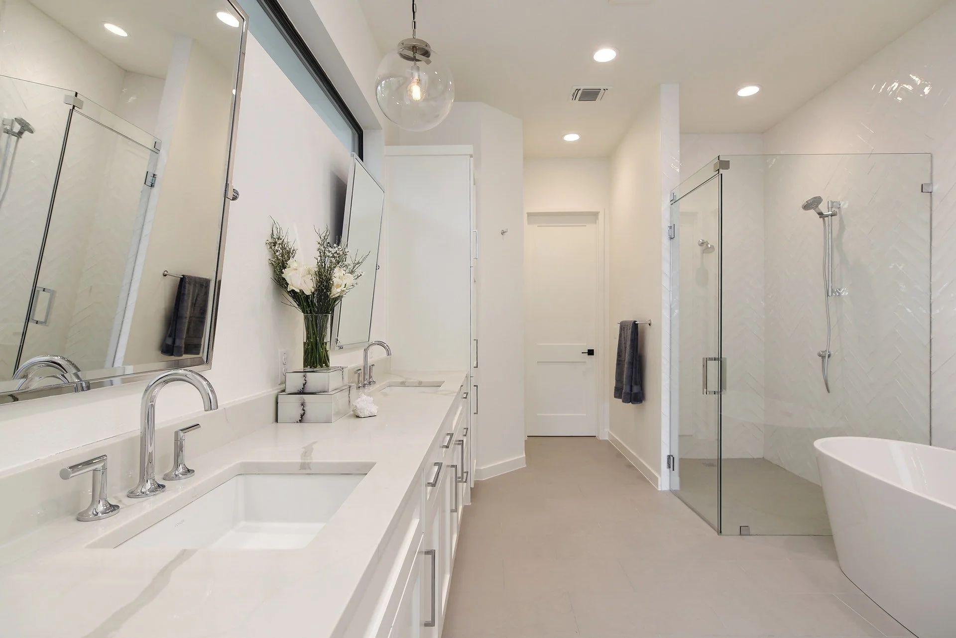 Modern white bathroom with double sinks, mirror, flowers, glass shower enclosure, and a free-standing bathtub.