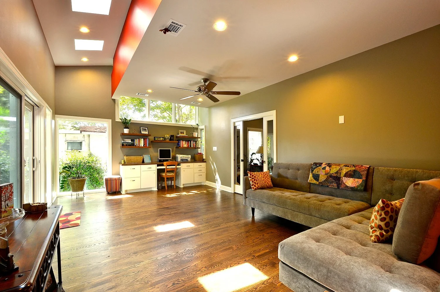 A spacious living room with hardwood floors, a large sliding glass door, a built-in desk and shelves, a ceiling fan, and a long gray sectional sofa with decorative pillows.