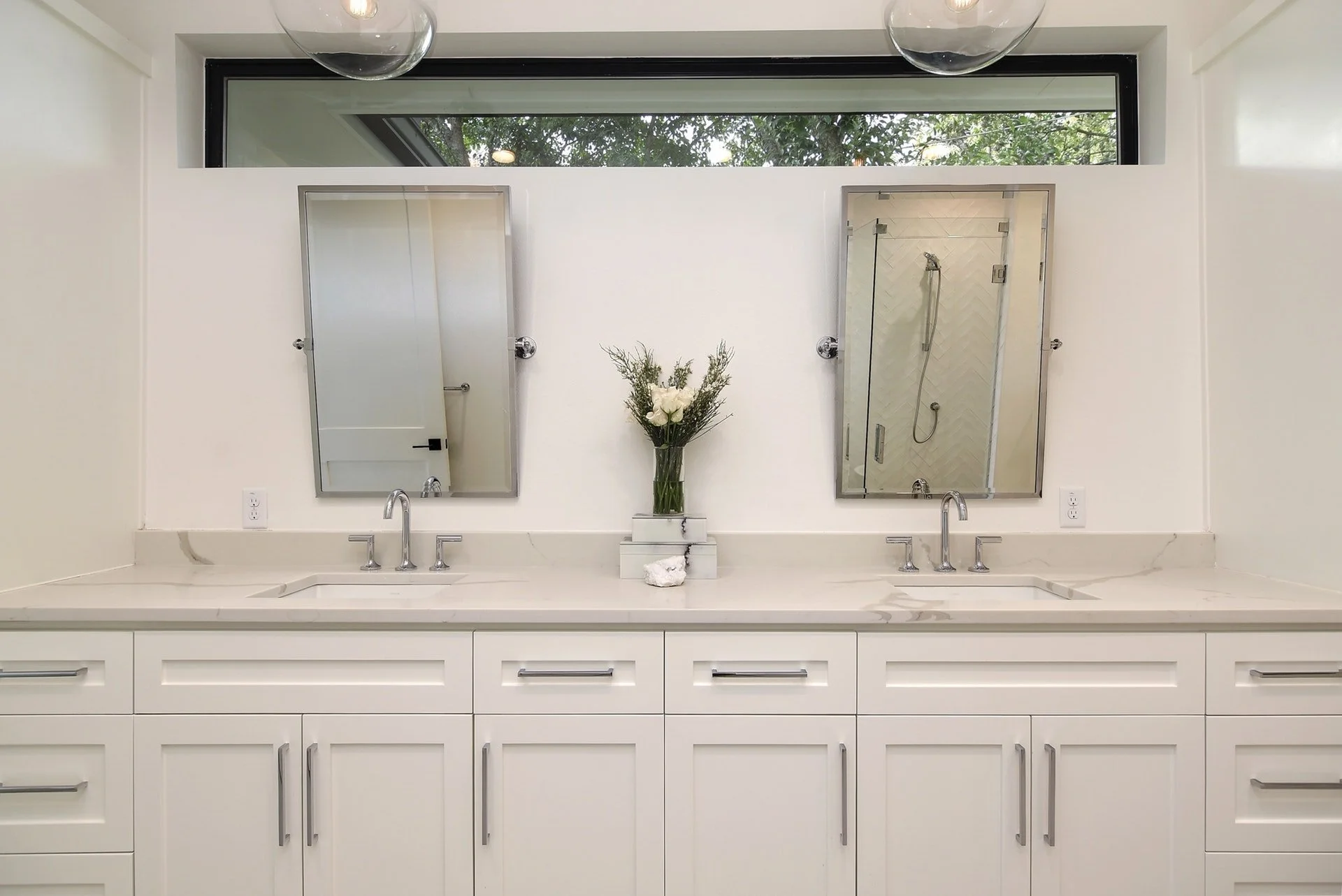 White bathroom vanity with two sinks, two mirrors, and a vase of flowers in the center. A window is above the mirrors, and there's a shower visible through a glass door in the background.