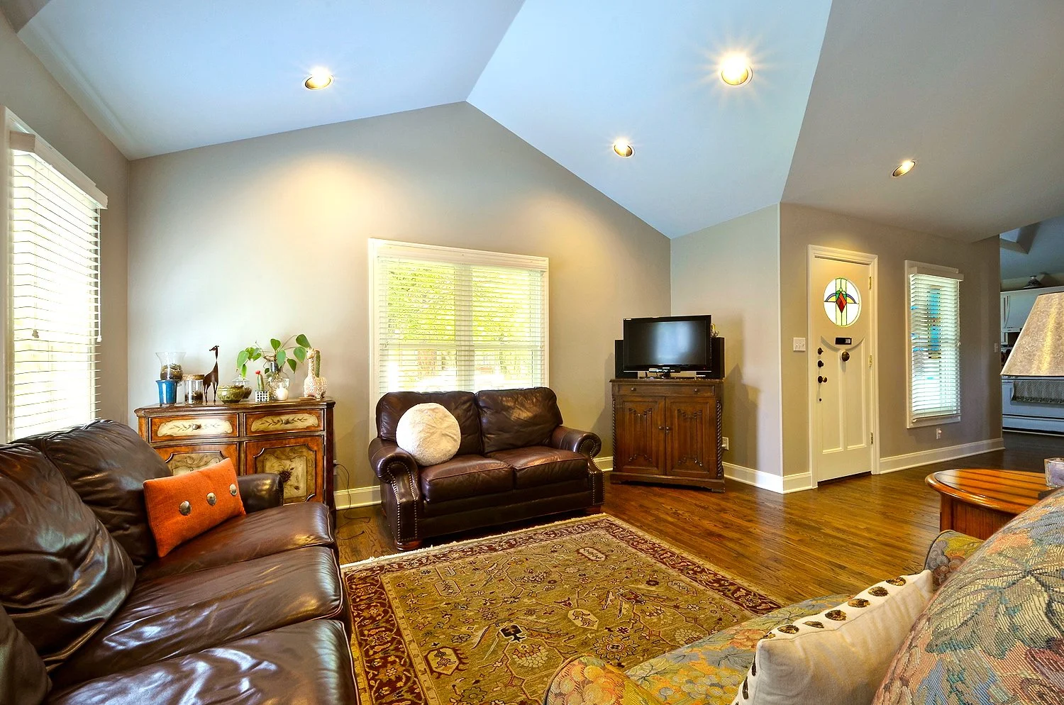 Living room with leather sofas, wooden flooring, a patterned area rug, potted plants, a wooden cabinet with decorations, a flat-screen TV, and windows with white blinds.