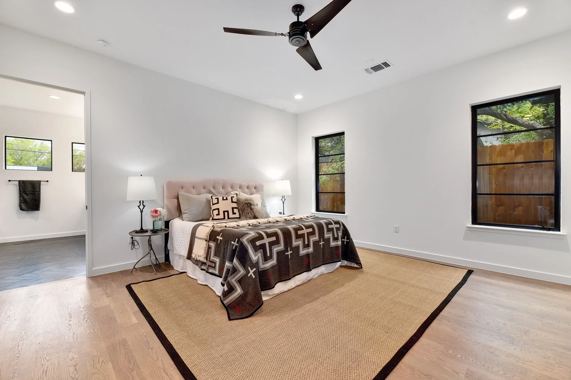 A bedroom with white walls, a bed with a black weather blanket, pillows, and a headboard. There are two nightstands with lamps, and windows with black frames showing trees outside. The room has wood flooring and a beige rug. A ceiling fan is mounted 