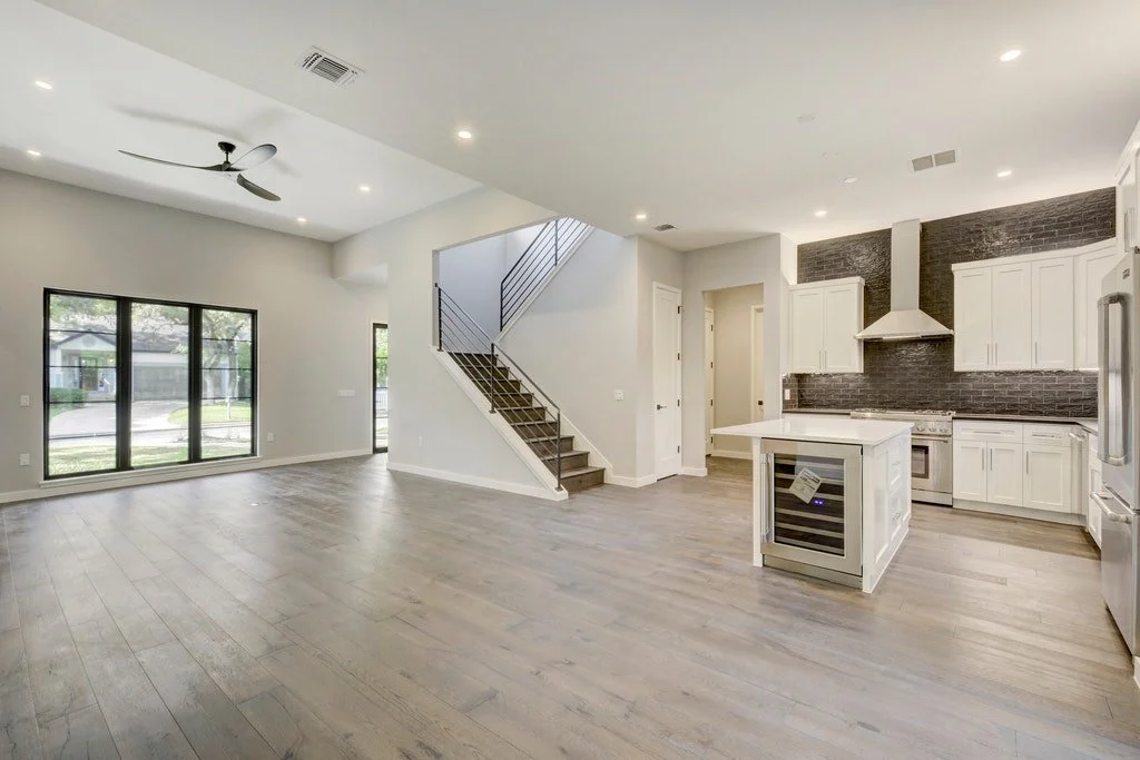 Open floor plan living area and kitchen in a modern home with white cabinets, dark brick backsplash, and kitchen island, with sunlight streaming through large windows.