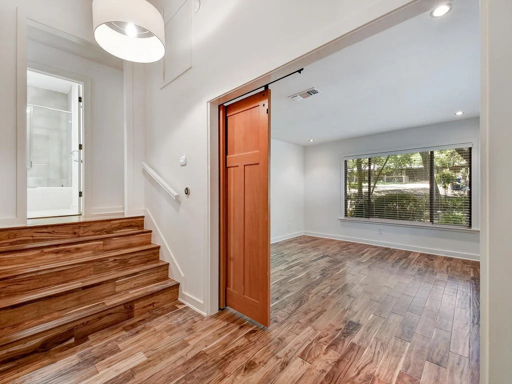 Interior view of a house showing a staircase, a room with wood flooring, a large window, and a sliding wooden door leading to the room.