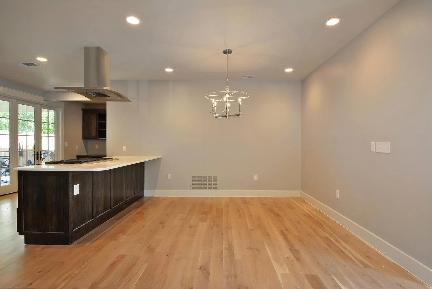 Empty dining area with hardwood flooring, a modern chandelier, and a part of the kitchen island visible on the left, with sliding glass doors leading outside.