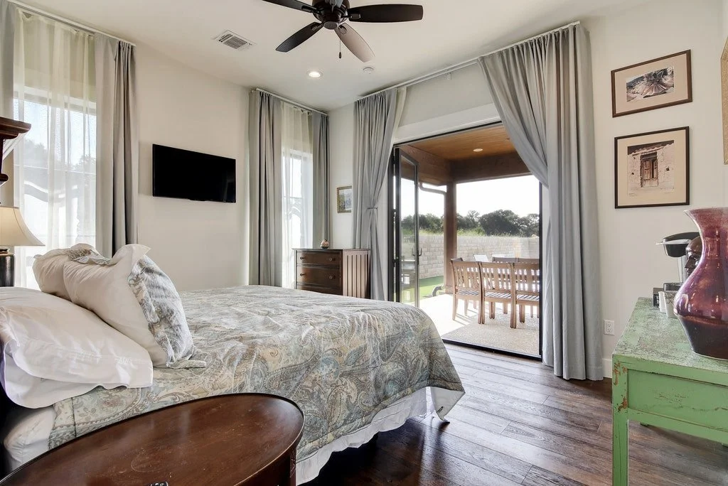 A cozy bedroom with a large bed covered in a light paisley-patterned blanket, a small wooden bedside table, a wall-mounted TV, and sliding glass doors leading to a balcony with outdoor seating overlooking a grassy landscape.