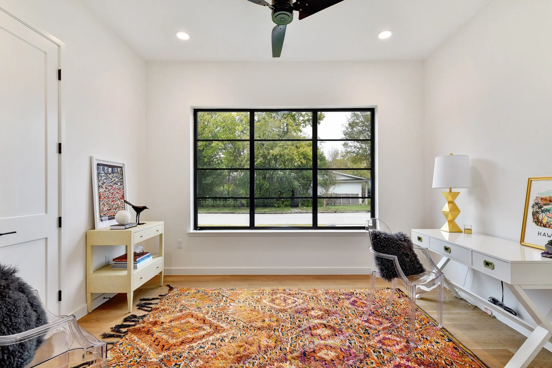 A bright, modern living room with a large window showing green trees outside. The room has white walls, a colorful rug, a white dresser with a gold lamp, a framed artwork, and a clear chair with a furry black pillow.
