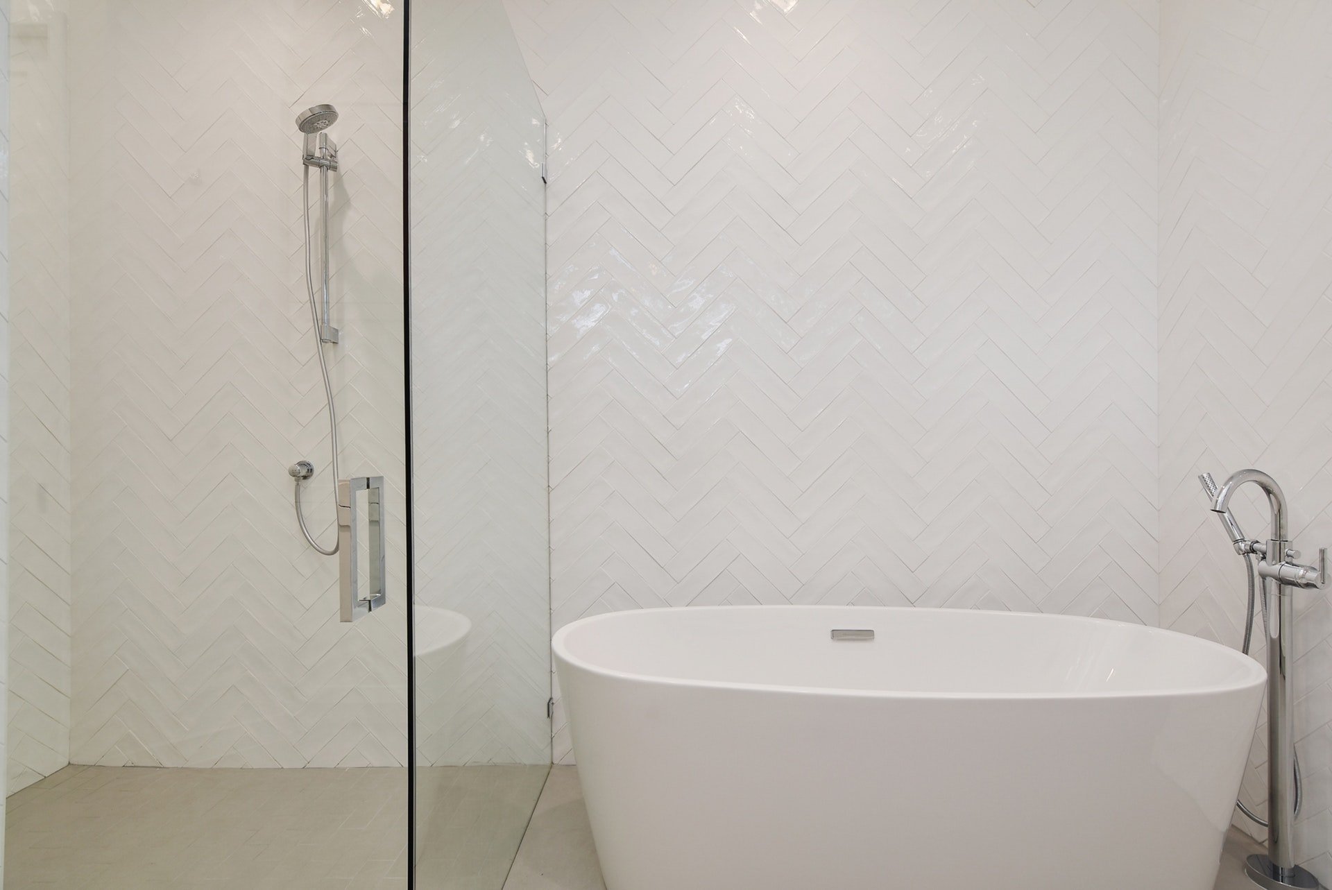 Modern minimalist bathroom with a white freestanding bathtub, a glass-enclosed shower area with a wall-mounted showerhead, and white herringbone-patterned tiled walls.