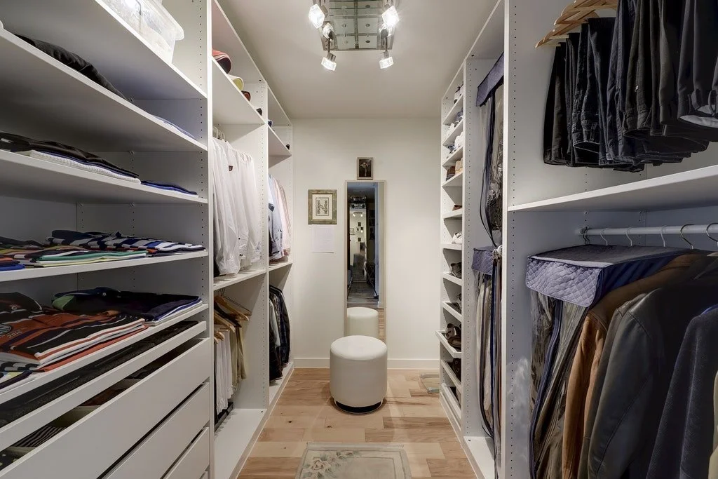 Walk-in closet with white shelves filled with folded clothes and hanging garments, a full-length mirror, and a white round ottoman in the center.