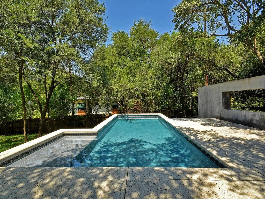 A backyard swimming pool with a concrete deck, surrounded by trees and greenery, under a clear blue sky.