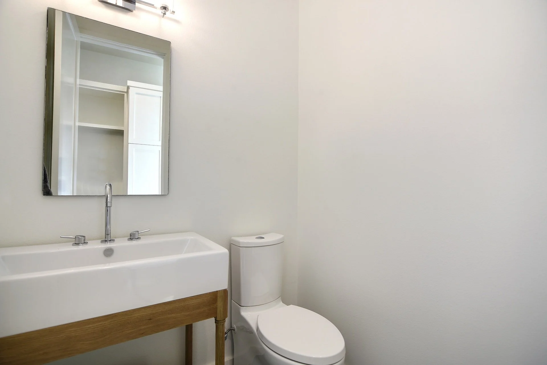 A minimalist bathroom with a white sink on a wooden vanity, a modern white toilet, a rectangular mirror above the sink, and white walls.