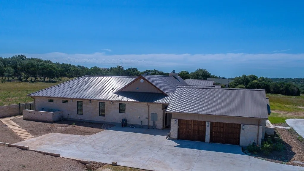 A modern house with a metal roof, concrete driveway, and surrounding yard, located in a rural area with trees and open land.