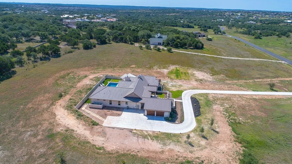 A large house under construction with a paved driveway, surrounded by open land and some trees, in a rural area with other houses in the distance.