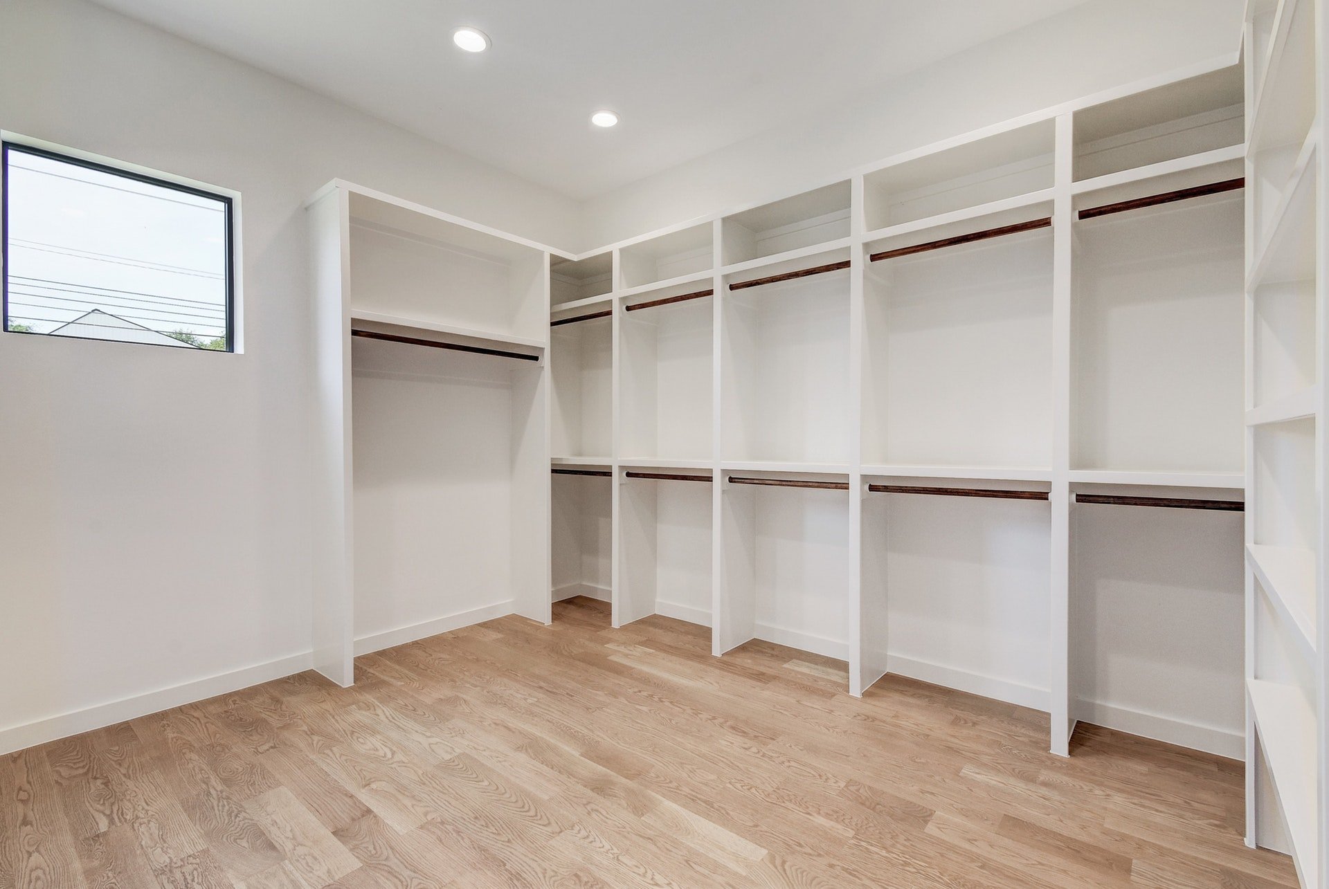 Empty walk-in closet with white shelving units and wooden rods, hardwood flooring, and a small window