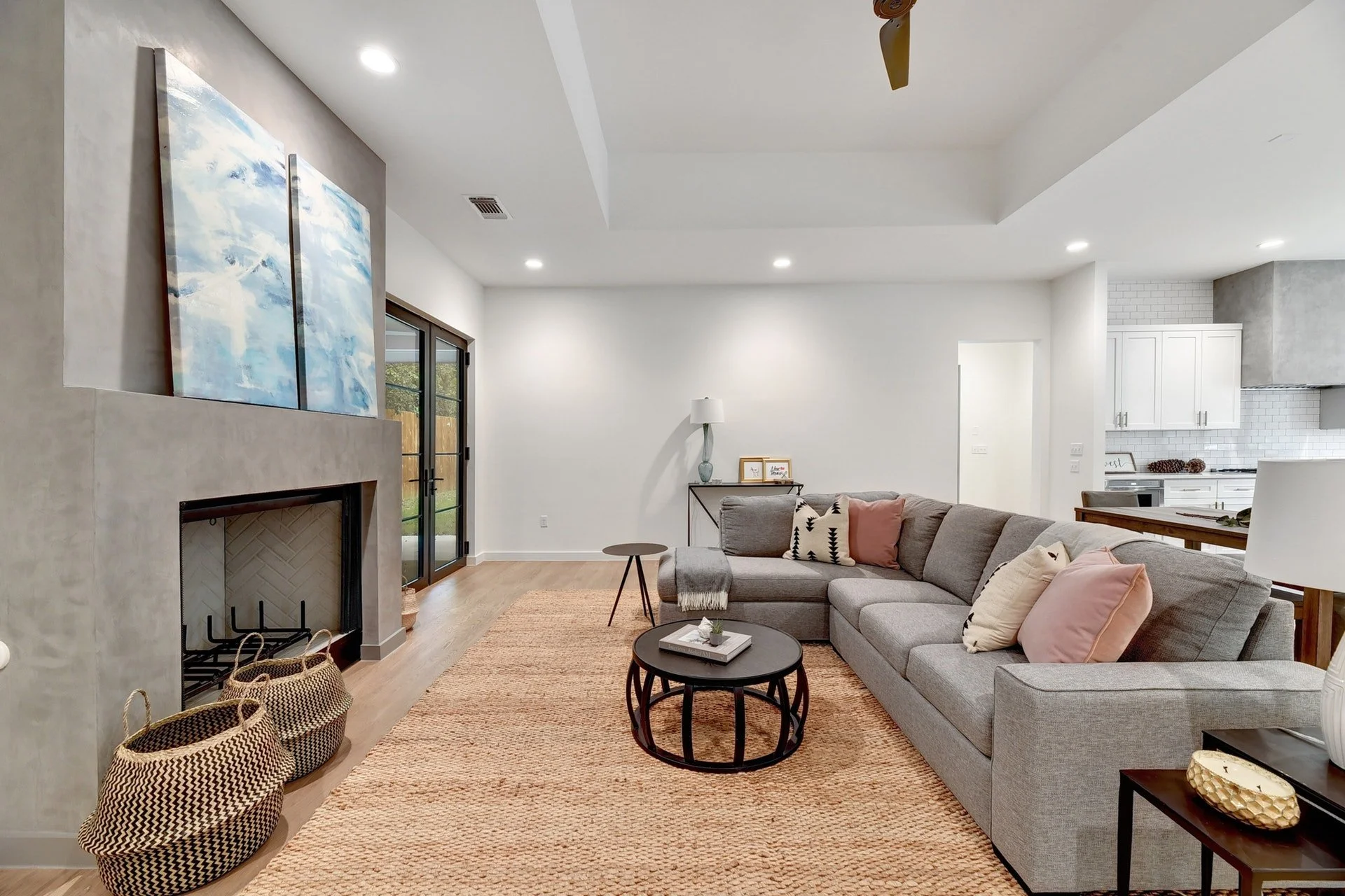 Living room with a large gray sectional sofa, round black coffee tables, and a woven area rug. Fireplace with a painting above it, a sliding glass door, and white kitchen cabinets in the background.