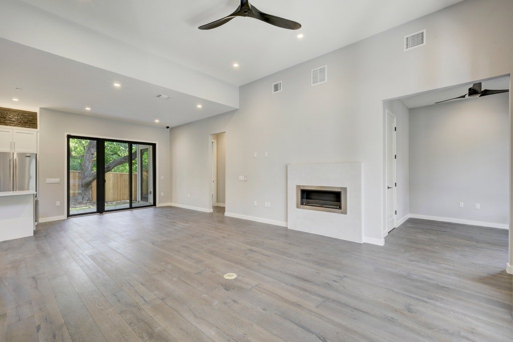 Empty living room with light gray walls, hardwood floors, a fireplace, ceiling fans, and sliding glass doors leading to an outdoor area.
