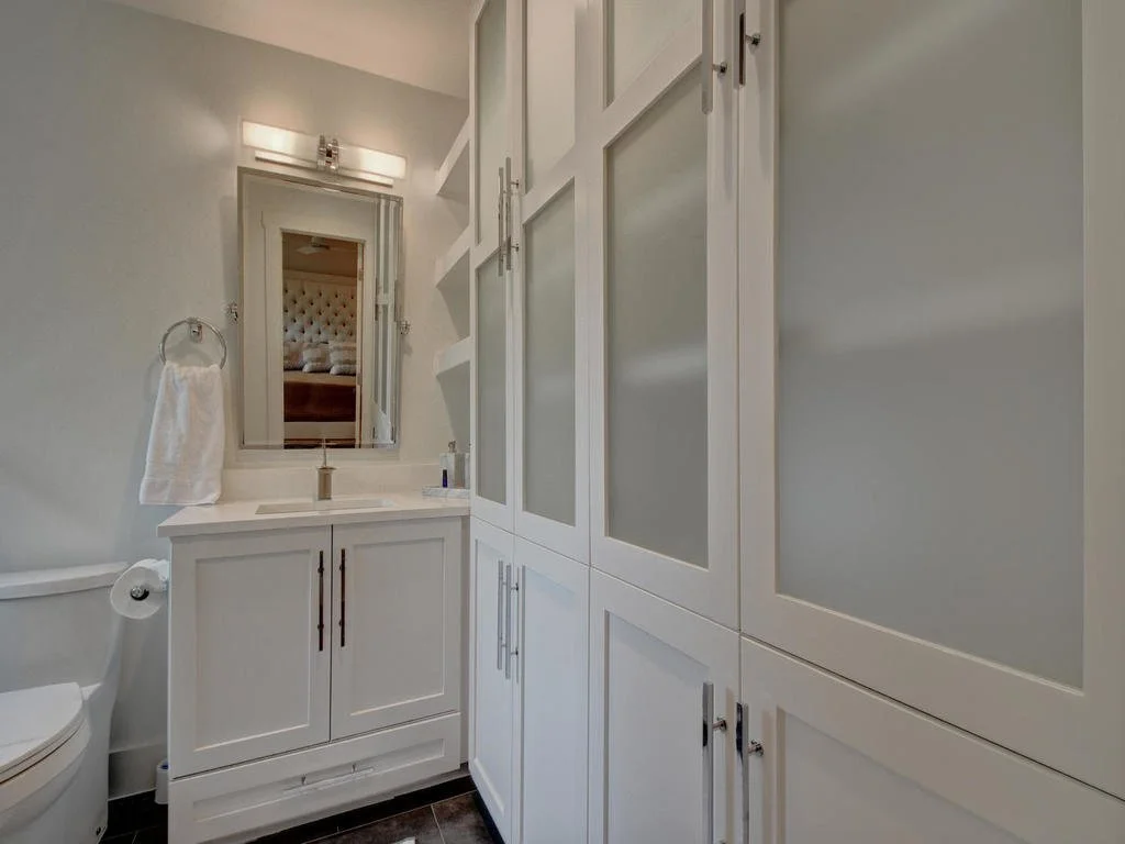 A small bathroom with a white vanity, mirror, and cabinets, a toilet, and a towel on a ring, with light gray walls and dark floor tiles.