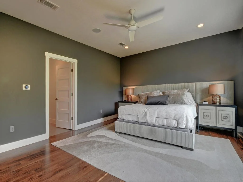 A bedroom with a bed against a green wall, two nightstands with lamps, a ceiling fan, hardwood floors, and a curved area rug.
