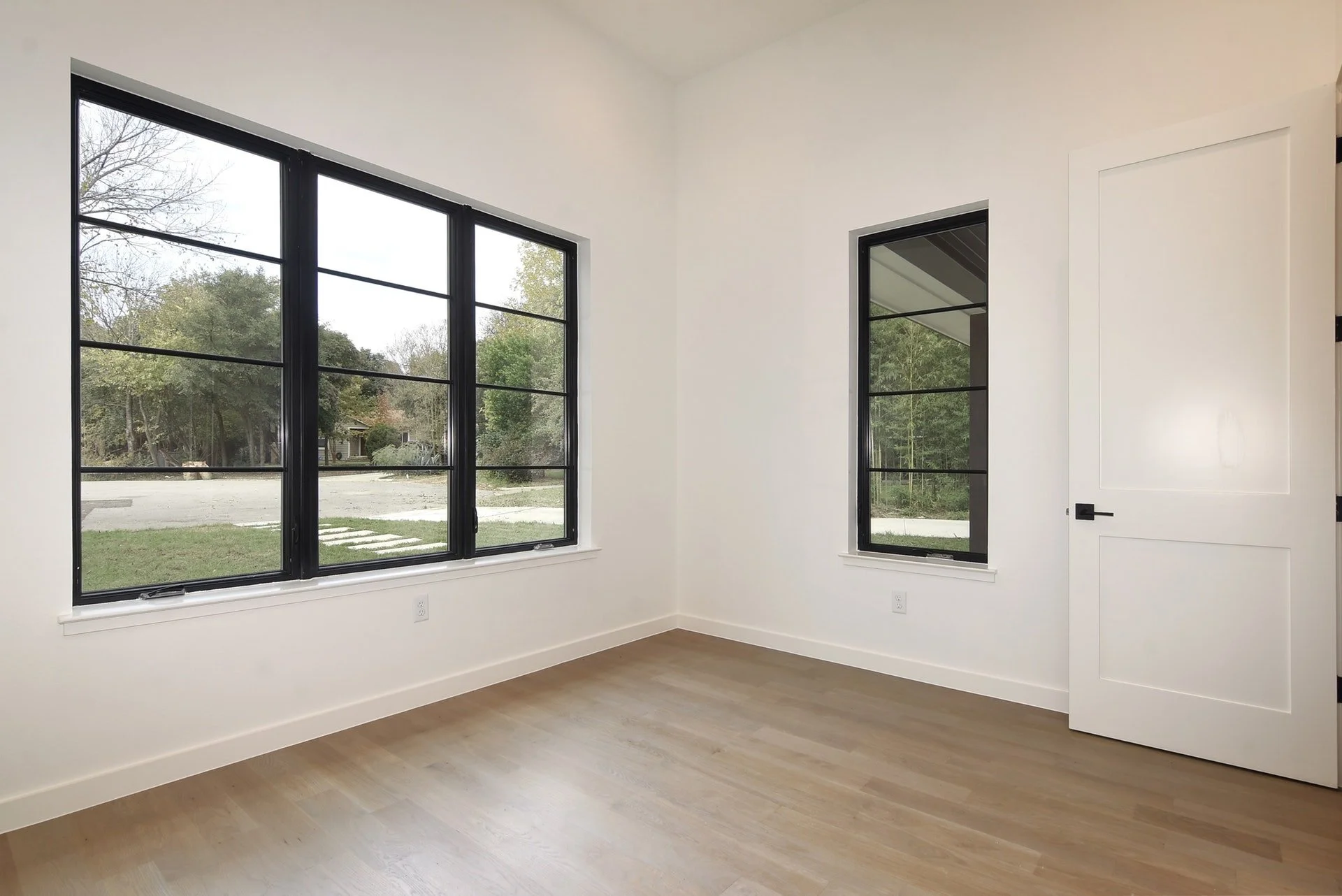 Empty room with white walls, light wood flooring, a large window and a smaller window showing trees outside, and a white door.